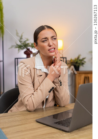 Young businesswoman touches cheek feels tooth pain while working on laptop and slows typing pace Young businesswoman touches cheek feels tooth pain while working on laptop and slows typing pace 130267113
