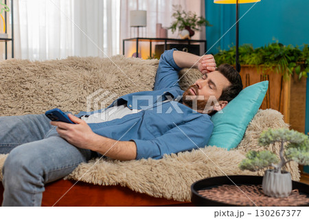 Young man unexpectedly falling asleep on sofa while holding smartphone in hand, tired and exhausted Young man unexpectedly falling asleep on sofa while holding smartphone in hand, tired and exhausted 130267377