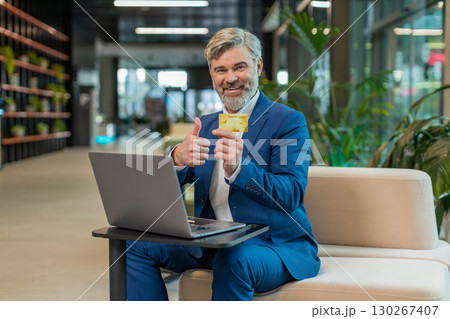 Smiling businessman using credit card and laptop for online shopping payments in modern office lobby Smiling businessman using credit card and laptop for online shopping payments in modern office lobby 130267407