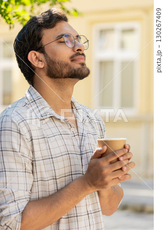 Happy Indian young man enjoying morning coffee hot drink and smiling standing on city street 130267409