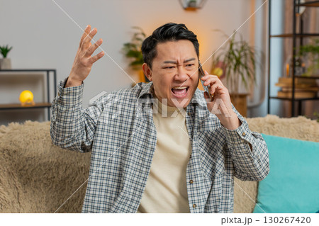 Angry young Chinese man arguing on smartphone phone while sitting on couch in living room at home 130267420