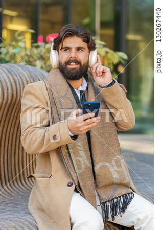 Happy Indian man in wireless headphones listening music in smartphone dancing sitting on bench Happy Indian man in wireless headphones listening music in smartphone dancing sitting on bench 130267440