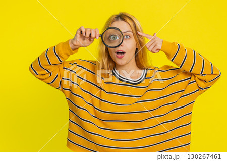 Young woman holds magnifying glass near face looking at camera with big zoomed eye, analyzing 130267461