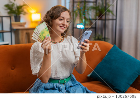Successful young woman holding smartphone and euro bill money fan sitting on sofa couch at home 130267608