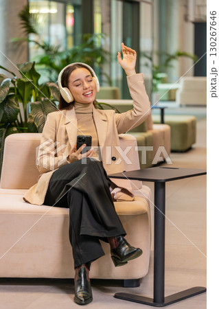 Excited young businesswoman in headphones listening energetic music on smartphone in office lobby Excited young businesswoman in headphones listening energetic music on smartphone in office lobby 130267646