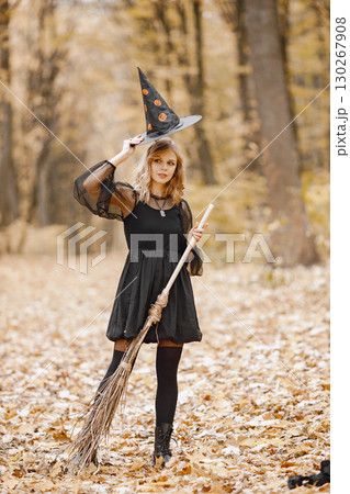Blonde young girl witch in forest on Halloween. Girl wearing black dress and cone hat. Witch holding a broom. 130267908