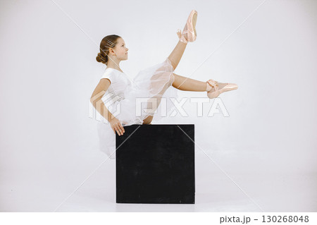 Little ballerina on white background. Brunette girl wearing white ballet suit. Girl sitting on black cube. 130268048