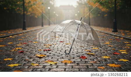 A clear umbrella lies discarded on a glistening cobblestone path, adorned with scattered colorful autumn leaves, set against a misty, tree-lined street scene. 130268211