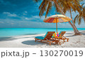 Two beach chairs and a parasol on the sand in a tropical beach. Summer vacation relaxing on white sand beach with palm trees. Oceanfront holiday scene with palm trees and blue sky. 130270019