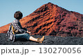 A girl sits on a black sand, enjoys a volcanic mountain landscape. 130270056