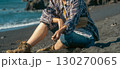 A girl in casual clothes sits on a black volcanic beach by the Atlantic ocean. 130270065