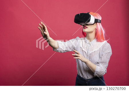 Curious young woman wearing virtual reality headset, raising one arm, navigating through futuristic 3D experience. Female model with pink hair, immersed in tech innovation and digital exploration. Curious young woman wearing virtual reality headset, raising one arm, navigating through futuristic 3D experience. Female model with pink hair, immersed in tech innovation and digital exploration. 130270700