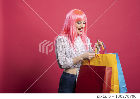 Gleeful young woman with pink wig smiling brightly and holding vibrant shopping bags. Stylish white lady in casual outfit posing happily after successful shopping spree, isolated background. Gleeful young woman with pink wig smiling brightly and holding vibrant shopping bags. Stylish white lady in casual outfit posing happily after successful shopping spree, isolated background. 130270706
