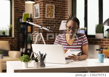 Young black woman prepares for project pitch, watching online course on laptop. Female freelancer seated at home office desk, writing in notebook, learning new skills through virtual training. 130270751