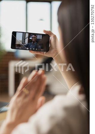 Young businesswoman doing hello hand gesture at black male colleague during online meeting on smartphone. Remote professionals collaborate on strategy during virtual call in home office. 130270792