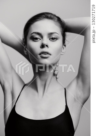 Elegant young woman in a black tank top posing gracefully, showcasing natural beauty and confidence against a soft gray background. Artistic black and white portrait. 130271719