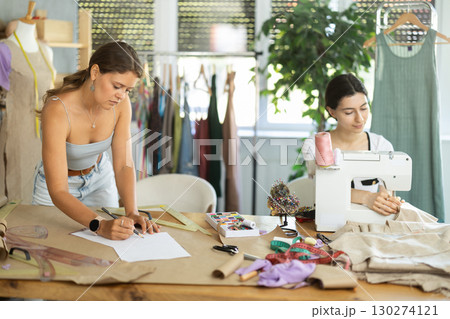 Female fashion designer sketching outfit while teammate sewing at machine Female fashion designer sketching outfit while teammate sewing at machine 130274121