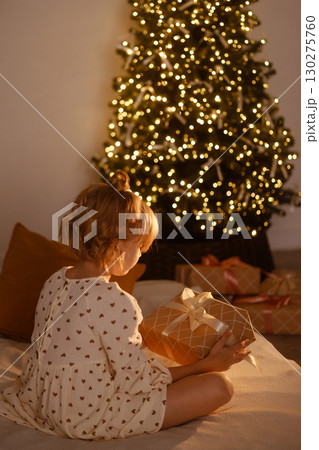 Young girl holding Christmas gift sitting on bed near decorated tree with lights, rear view 130275760