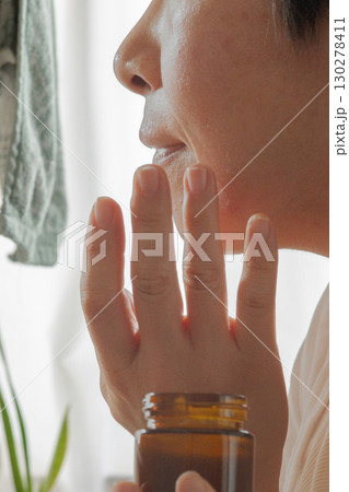 鏡に向かって赤みがかった頬にローションを塗る女性の横顔の下半分が移った鏡 130278411