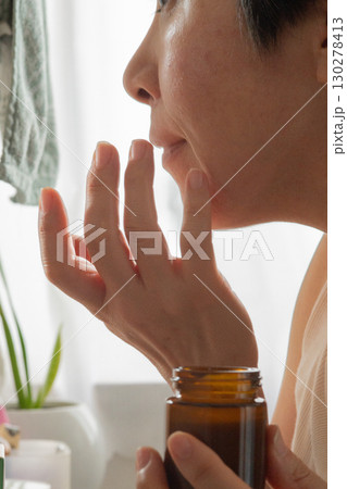 鏡に向かって赤みがかった頬にローションを塗る女性の横顔の下半分が移った鏡 130278413