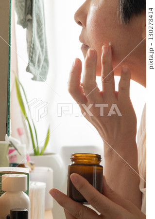 鏡に向かって赤みがかった頬にローションを塗る女性の横顔の下半分が移った鏡 130278414