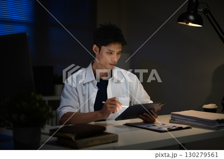 Focused businessman working late at night, analyzing documents on digital tablet in dark office Focused businessman working late at night, analyzing documents on digital tablet in dark office 130279561