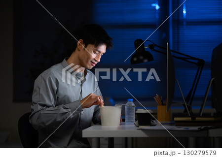 Office worker eating instant noodles while working overtime in dimly lit office 130279579