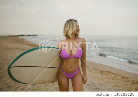 Attractive young woman in a purple bikini posing with a surfboard on the beach, enjoying summer by the sea, confident and relaxed, ocean waves in the background. Attractive young woman in a purple bikini posing with a surfboard on the beach, enjoying summer by the sea, confident and relaxed, ocean waves in the background. 130282220