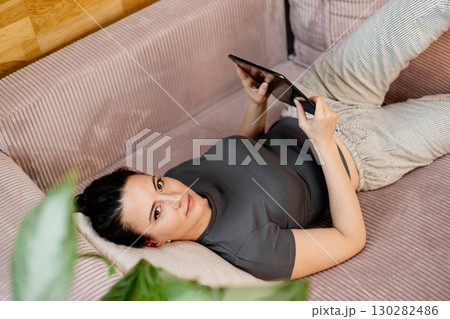 Relaxed woman taking a break, lying on sofa and scrolling on tablet. Relaxed woman taking a break, lying on sofa and scrolling on tablet. 130282486