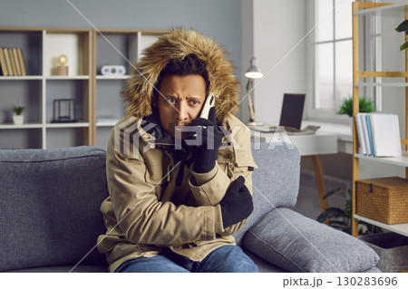 Young frozen man sitting in a winter coat on the sofa at home and talking on the phone. Young frozen man sitting in a winter coat on the sofa at home and talking on the phone. 130283696