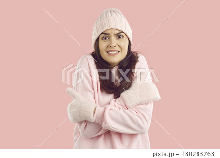 Woman freezes in pink sweatshirt, mittens and hat hugging herself by hands on pink background. Woman freezes in pink sweatshirt, mittens and hat hugging herself by hands on pink background. 130283763