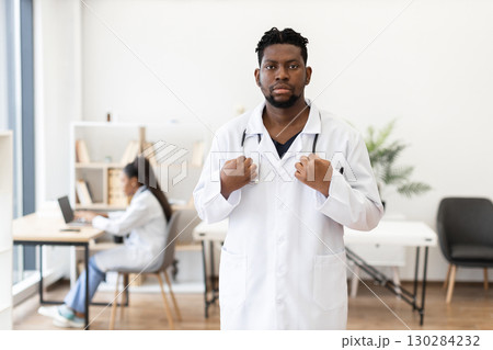 Confident male healthcare professional, adult, in white coat standing in modern medical workspace. Bright and spacious clinic setting with a colleague working in the background. 130284232