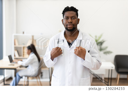 Confident male healthcare professional, adult, in white coat standing in modern medical workspace. Bright and spacious clinic setting with a colleague working in the background. Confident male healthcare professional, adult, in white coat standing in modern medical workspace. Bright and spacious clinic setting with a colleague working in the background. 130284233