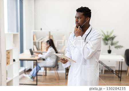 Male African doctor in early 30s using tablet in office, colleague working nearby, healthcare setting. Male African doctor in early 30s using tablet in office, colleague working nearby, healthcare setting. 130284235