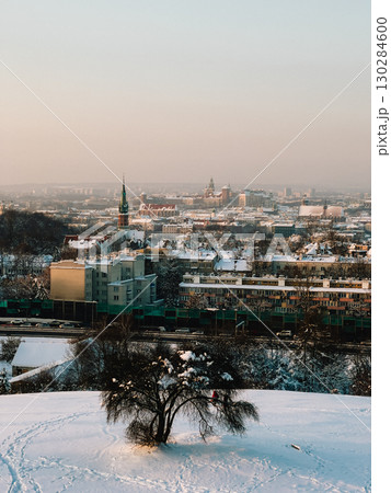 Panoramic view capturing enchanting cityscape of Krakow, blanketed in snow during stunning sunset. Wawel Castle stands majestically, framed by snowy tree in foreground. Snowy panorama, Krakow's winter 130284600