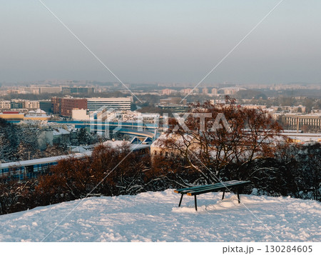 Empty bench sits on snow covered hill, overlooking cityscape of Krakow, Poland, during serene winter sunset, with light dusting of snow on trees and rooftops. Snow covered hill revealing wooden bench 130284605