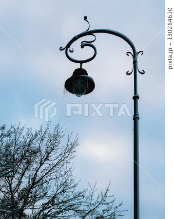 Ornate street lamp adorned with icicles hangs gracefully on cloudy winter day, while bare tree branches create serene silhouette in foreground, enhancing tranquil atmosphere. Street lamp with icicles 130284610
