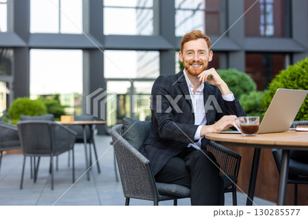 Smiling businessman with ginger hair sitting at urban cafe with laptop Smiling businessman with ginger hair sitting at urban cafe with laptop 130285577