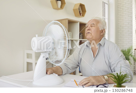 Senior man, tired of summer heat, trying to cool down by an electric fan at home 130286748