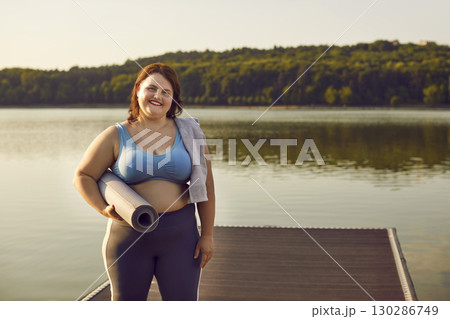 Smiling overweight woman in sportswear holding yoga mat and standing on lake pier Smiling overweight woman in sportswear holding yoga mat and standing on lake pier 130286749