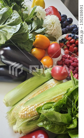 A colorful variety of fresh fruits and vegetables including corn, berries, tomatoes, eggplants and greens arranged on a white surface 130288691