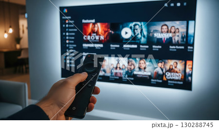 Hand with remote control in front of wall mounted smart TV with led backlighting, person choosing movie on streaming service in modern home theater 130288745