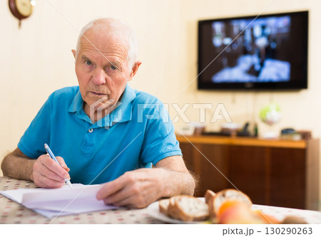 Serious older man writing letter at table in living room Serious older man writing letter at table in living room 130290263