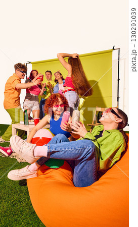Group of young friends laughing and drinking on beanbags with green backdrop 130291039