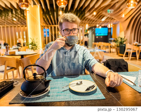 Man sipping hot tea at restaurant table with modern interior and warm lighting. Lifestyle, relaxation, leisure, and hospitality concept with cozy atmosphere and enjoyment. Man sipping hot tea at restaurant table with modern interior and warm lighting. Lifestyle, relaxation, leisure, and hospitality concept with cozy atmosphere and enjoyment. 130295067