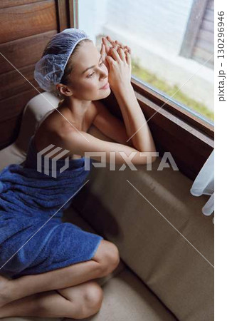 Relaxed woman wrapped in blue towel wearing shower cap, sitting by window with natural light, enjoying peaceful moment indoors in cozy wooden interior. 130296946