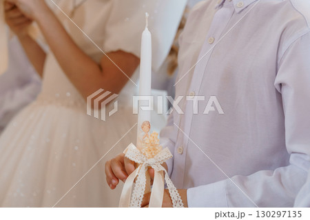 Orthodox Christian Baptism Ceremony: Young Boy with Lit Candle, White Garments, Lace Bow, Religious Ritual, Light Purple Background, Soft Focus. 130297135