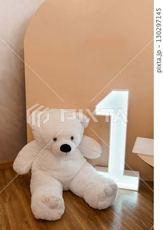 Adorable Giant White Teddy Bear Sitting Next to a Lit Number One, Celebrating First Birthday in a Pastel Pink Room. Adorable Giant White Teddy Bear Sitting Next to a Lit Number One, Celebrating First Birthday in a Pastel Pink Room. 130297145