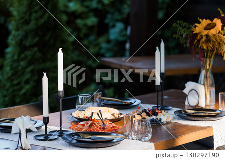 Outdoor Dinner Setting with Lit Candles, Black Plates and Charcuterie, Rustic Wooden Table, Lush Greenery, Evening Mood 130297190