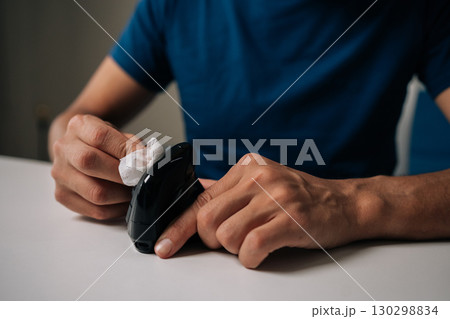 Close-up cropped shot of man cleaning modern black vertical mouse with disinfectant wipe, promoting hygiene and preventing spread of germs, sitting at white table in workplace or home office. 130298834